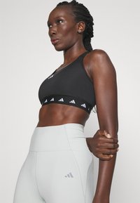 Sujetador deportivo negro con una textura suave y una banda elástica con marca; combinado con mallas deportivas de talle alto en gris claro.