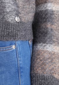 Neulottu harmaa villapaita beigeillä yksityiskohdilla, jossa on teksturoitu pinta ja metallinen nappi. Yhdistetty sinisiin farkkuihin, korostaen vyötärönauhaa.