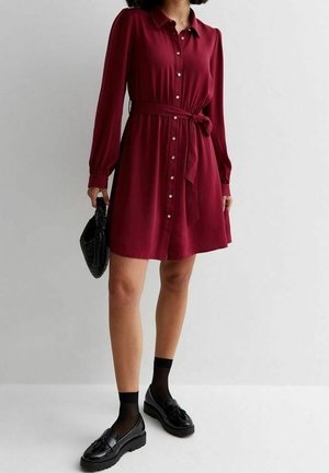 Bordeauxrode jurk met knopen, lange mouwen, ceintuur in de taille en soepelvallende stof. Gecombineerd met zwarte schoenen en een gestructureerde zwarte handtas.