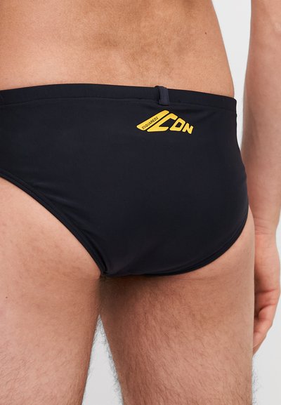 Shorts de bain noirs avec une texture lisse, comportant un logo jaune à l'arrière. Le design inclut une taille mi-haute et une couverture minimale.