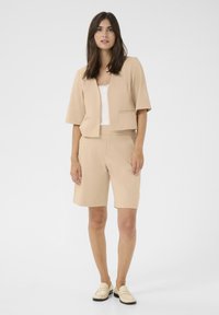 Beige bleiseri, jolla on lyhyet hihat, yhdistettynä saman värisiin shortseihin. Alla on teksturoitu valkoinen toppi, jota täydentävät vaaleanväriset loaferit.