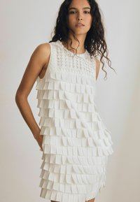 Valge kleit, millel on kihilised, ristkülikukujulised voldid ja tekstureeritud, tikitud kaelus. Kleit on ilma varrukateta ja sirge lõikega.