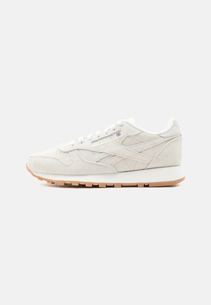 Classic low-cut white suede sneaker with textured sole, lace-up front, and subtle brand logo on side panel against plain background.