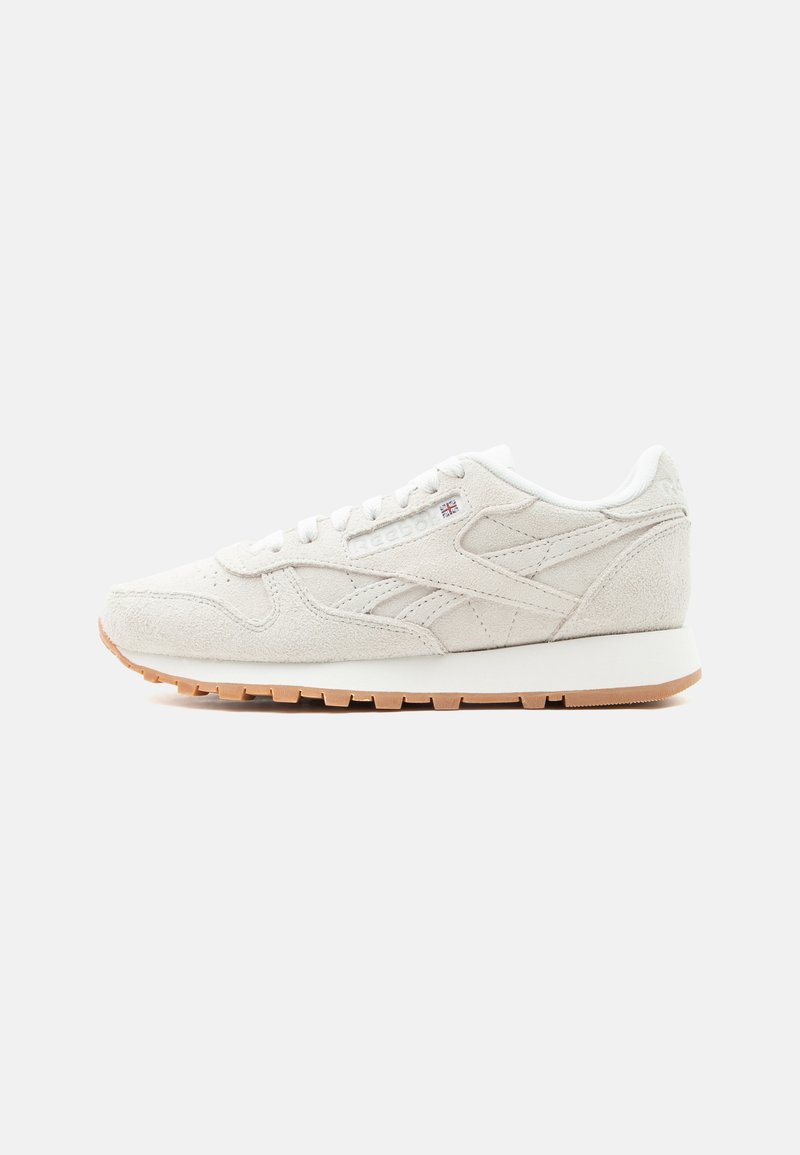 Κλασικό λευκό σουέντ αθλητικό παπούτσι με χαμηλή κοπή, υφή στη σόλα, δέσιμο με κορδόνια μπροστά και διακριτικό λογότυπο της μάρκας στην πλαϊνή επιφάνεια, σε απλό φόντο.