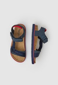 Jūriniai mėlyni sandalai su kamštiniu padu, velcro diržais ir raudonais akcentais ant pado ir nugaros. Audinė padas geresniam sukibimui. Matoma prekės ženklo etiketė.
