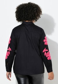 Schwarzes Hemd mit langen Ärmeln, das auf beiden Ärmeln leuchtend pinke Blumenstickerei aufweist. Der Stoff wirkt glatt und hat ein klassisches Kragen-Design.