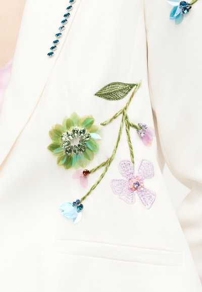 Λευκό ύφασμα με πράσινους κεντημένους βλαστούς και φύλλα, διακοσμημένο με μπλε, μοβ και ροζ παγέτες και χάντρες σε σχέδια λουλουδιών.