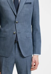 Mann trägt einen blauen, strukturierten Zweiknopf-Anzug mit Kerb- Revers, Pattentaschen, marineblauer Krawatte, weißem Hemd und passenden Anzughosen vor weißem Hintergrund.