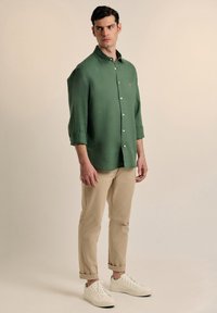 Camisa verde de botones con un corte relajado, mangas largas y puños enrollados, combinada con chinos beige y zapatillas blancas.