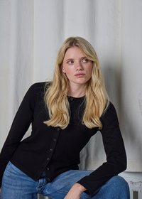 Cardigan nero a coste con scollo rotondo e chiusura frontale con bottoni, abbinato a denim blu. Tessuto morbido e design aderente. Sfondo chiaro.