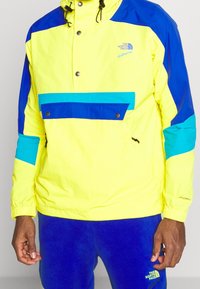 Giacca anorak giallo brillante con accenti blu, caratterizzata da una tasca frontale con bottoni a pressione, polsini elasticizzati e cappuccio regolabile per una vestibilità sicura.