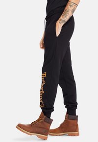 Pantaloni da jogging neri con tasche laterali, caratterizzati da un logo dorato "Timberland" sulla gamba sinistra. Abbinati a stivali marroni con lacci.