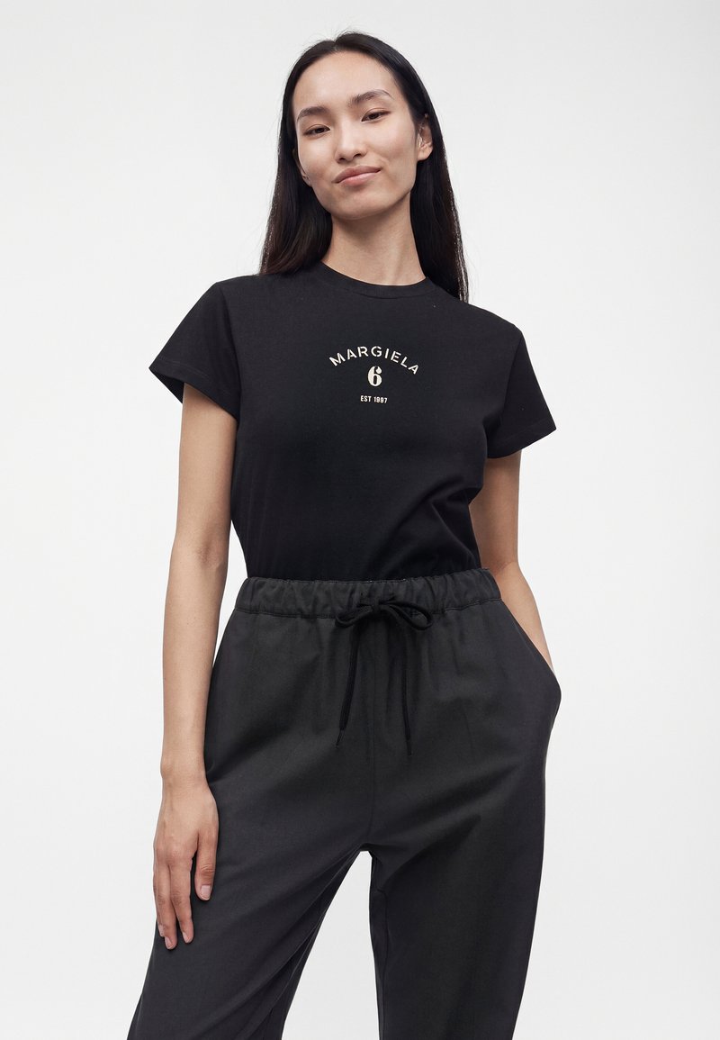 Femme portant un t-shirt noir de la marque Margiela et un pantalon noir à cordon, debout avec une main dans la poche sur fond blanc.