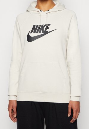 Persoon draagt een witte Nike-hoodie met zwart Swoosh-logo en zwarte trekkoorden, gecombineerd met losse zwarte broek, staand tegen een effen achtergrond.