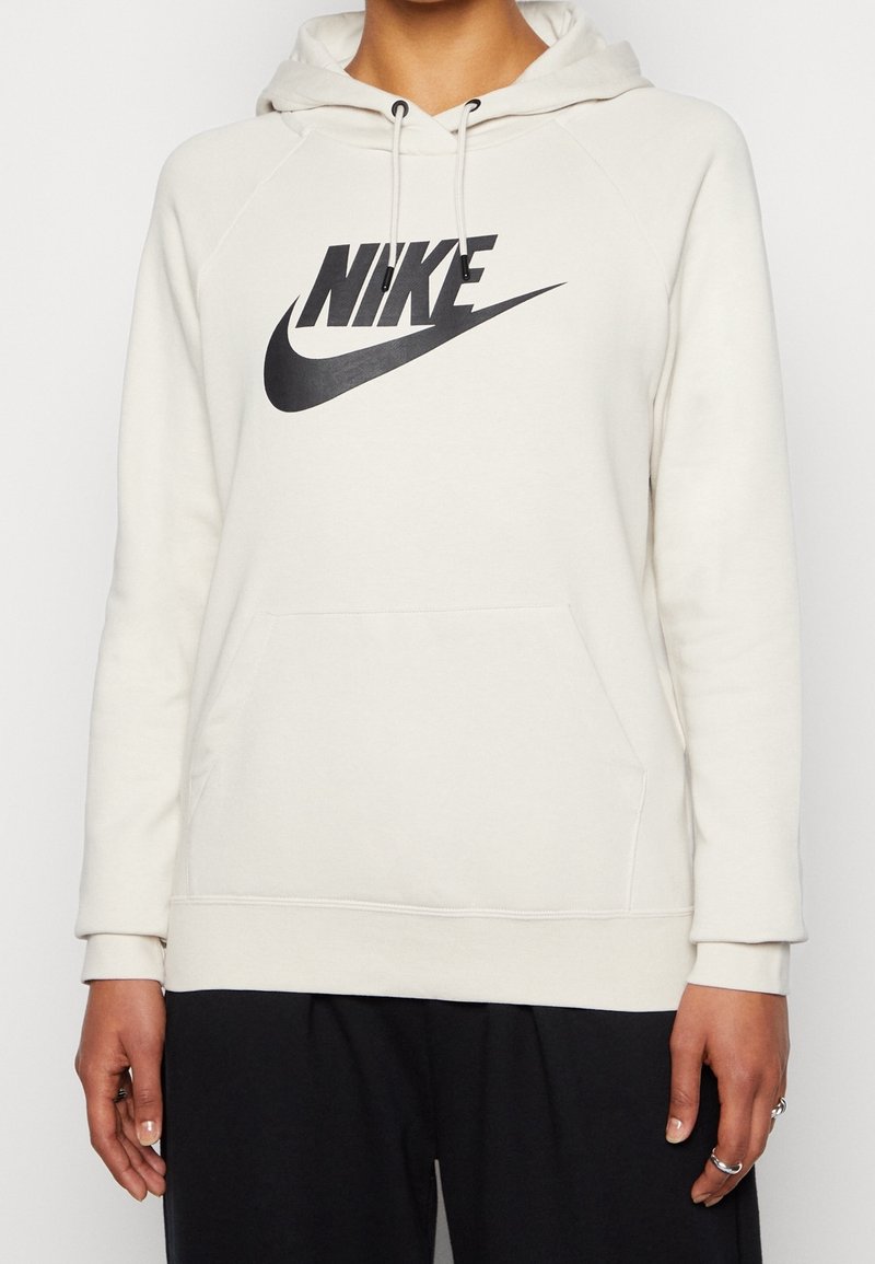 Personne portant un sweat à capuche blanc Nike avec un logo Swoosh noir et des cordons noirs, assorti à un pantalon noir ample, debout devant un fond uni.