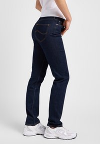 Persoon die donkerblauwe jeans met rechte pijpen en witte sneakers draagt, staand met één hand in de zak tegen een effen achtergrond.