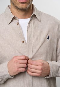Camisa de botones a rayas beige, hecha de una tela ligera, con un pequeño logo bordado, botones de madera y mangas largas.