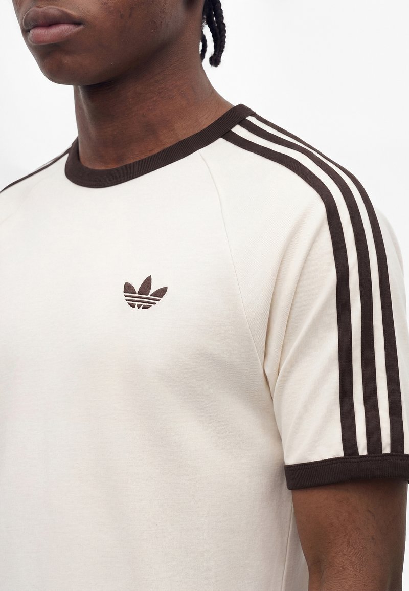 Junge Person trägt ein cremefarbenes Adidas T-Shirt mit dunkelbraunem Besatz und drei Streifen an den Ärmeln, mit Adidas Trefoil-Logo auf der Brust.