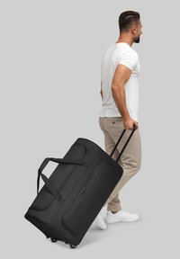 Schwarze Radreisetasche aus strukturiertem Stoff, mit doppelten Seitengriffen und zwei glatten, ausziehbaren Rädern. Offene Reißverschlusstasche auf der Vorderseite.