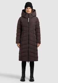 Mørkebrun polstret parka med hætte, har en lynlås i fuld længde, chevronmønster og er kombineret med sorte leggings og kraftige sorte støvler.