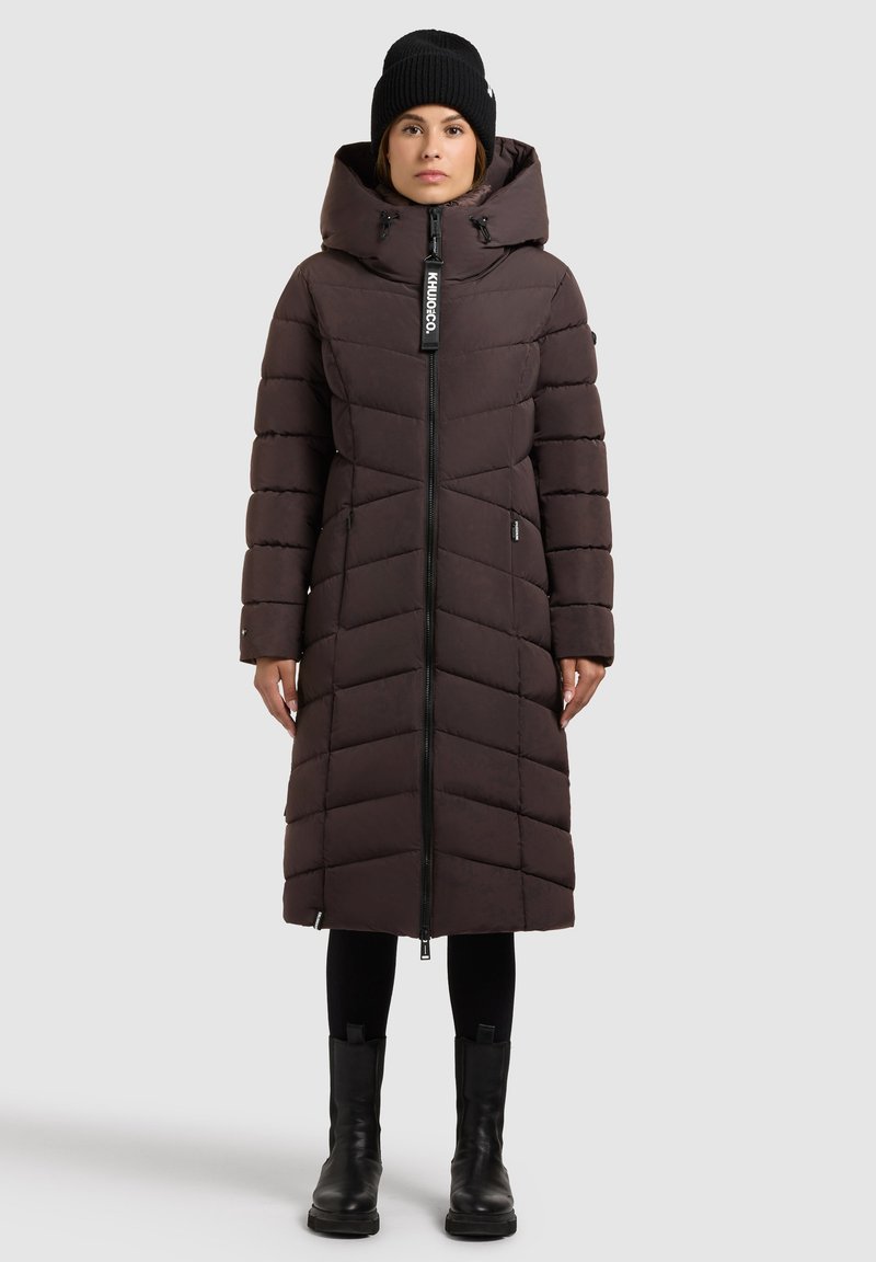 Mørkebrun polstret parka med hætte, har en lynlås i fuld længde, chevronmønster og er kombineret med sorte leggings og kraftige sorte støvler.