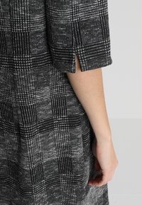 Abito a fantasia check bianco e nero che mostra una manica ripiegata con uno spacco laterale. Il tessuto appare texture e tessuto con una forma strutturata.