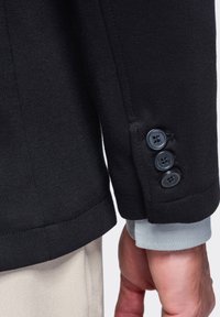 Gros plan d'une manche de blazer noir avec trois boutons noirs sur un poignet de chemise bleu clair et un pantalon beige, montrant une partie du bras d'un homme.
