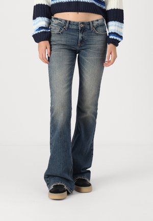 Femme portant un jean bootcut bleu délavé, des chaussures noires avec semelles beige, et un pull court en maille à rayures bleues, blanches et marine.