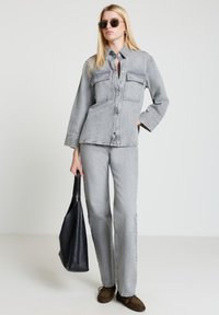 Graues Jeanshemd mit zwei Brusttaschen, kombiniert mit passenden grauen Jeans. Das Model hält eine schwarze Leder-Tasche und trägt eine dunkle Sonnenbrille.