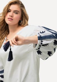 Top blanco con bordado floral azul y acentos de encaje, con mangas abullonadas y detalle de borlas en el escote. Textura suave.
