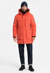 Heldere oranje geïsoleerde parka met een capuchon, voorzien van meerdere voorzakken, reflecterende accenten en een relaxte pasvorm. Zwarte jeans en laarzen.