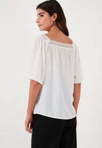 White blouse with short, wide sleeves and a scooped neckline. Features a lace trim along the neckline. Soft, lightweight fabric, slightly textured.