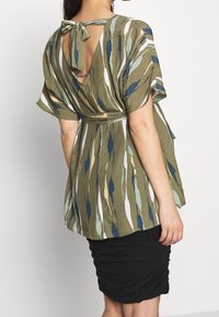 Olive green top with abstract patterns in blue, white, and beige. Features a tie-back design and an open back with an accent detail.