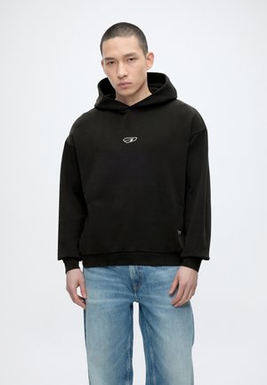 Ung mand iført en sort hættetrøje med en lille hvid logo og lyseblå jeans, stående mod en ensfarvet hvid baggrund.