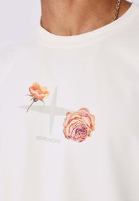 Camiseta blanca con rosas naranjas bordadas y un sutil logo "PXP" en gris, cuello redondo, tejido suave y textura lisa.