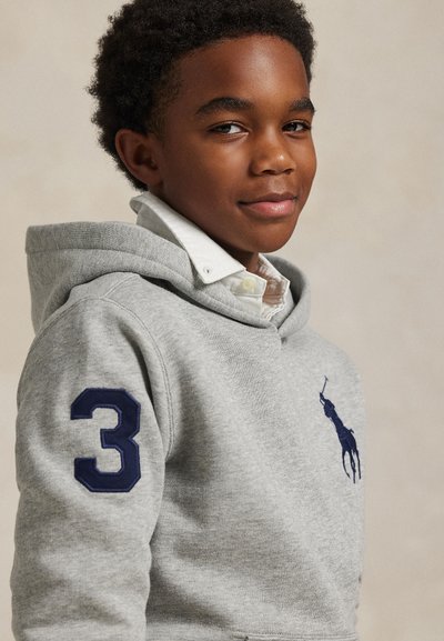 Garçon portant un sweat à capuche gris avec un logo joueur de polo bleu marine et un écusson numéro trois sur la manche, souriant légèrement.