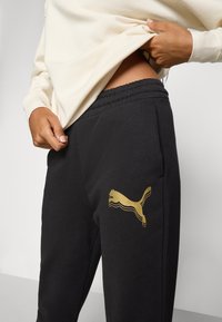 Sorte joggebukser med en gullfarget Puma-logo på venstre bein, med elastisk liv og mykt, teksturert stoff.