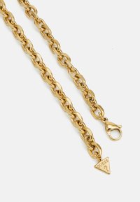 Gouden metalen ketting met ovale schakels, voorzien van een musketonhaak en een driehoekig label met de tekst "GUESS". Glad oppervlak en glanzende afwerking.