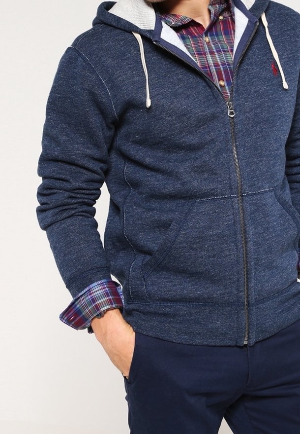 Marineblauwe zip-up hoodie gemaakt van gestructureerde stof, met een contrasterende lichtgrijze binnenvoering en kangoeroezakken, gedragen over een geruit overhemd.