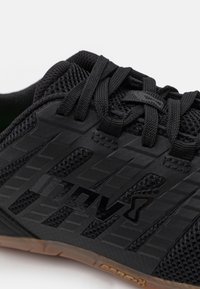 Close-up van een zwarte sportieve schoen met mesh-stof, gestructureerde synthetische overlays, zwarte veters en een gomrubberen zool.