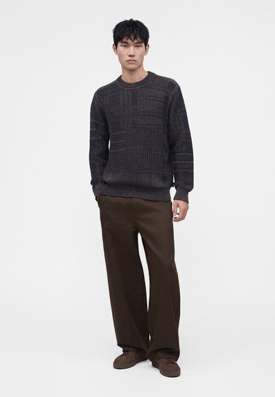 Homme portant un pull en maille texturée foncée, un pantalon large marron et des baskets marron, debout devant un fond clair uni.