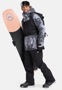 Mann in Wintersportbekleidung, der ein zweifarbige Snowboard hält und eine gemusterte Jacke, schwarze Hose, Handschuhe, eine Mütze, Skibrille und einen Rucksack trägt.