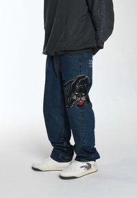 Jeans in denim con ricamo di una pantera nera sulla gamba sinistra, vestibilità relax, colore blu, indossati con sneakers bianche e una felpa scura.