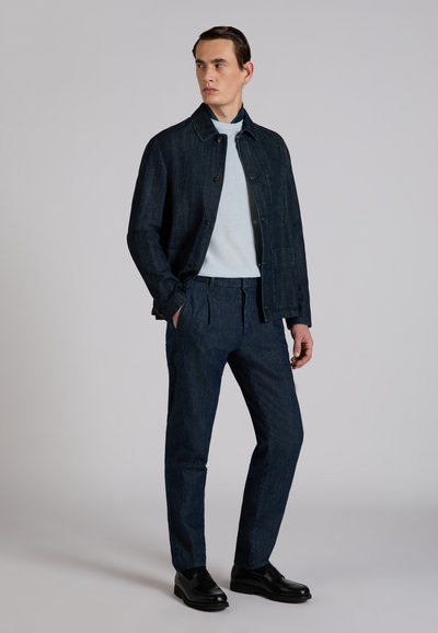 Mężczyzna stojący, ubrany w ciemną kurtkę i spodnie z denimu, jasnoniebieski sweter z golfem oraz czarne eleganckie buty, z rękami w kieszeniach, patrzący w lewo.