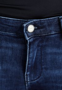 Vaqueros de mezclilla azul oscuro con un botón plateado, que presentan una textura suave y un diseño clásico de cinco bolsillos. Talle medio y trabillas para el cinturón incluidas.