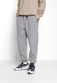 Graue Jogginghosen mit elastischen Bündchen, versehen mit einem schwarzen Logo am linken Oberschenkel. Kombiniert mit mehrfarbigen Sportschuhen.