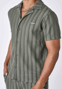 Camicia a maniche corte con bottoni di colore verde oliva, con righe verticali bianche, tessuto testurizzato, colletto e taschino sul petto con un accento argentato.