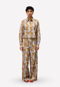Femme debout portant une chemise à manches longues à motif marron et un pantalon large, associé à des chaussures rouges, sur un fond blanc uni.