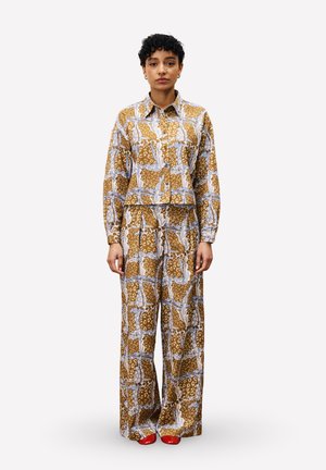 Vrouw staat in een bruine, patterned blouse met lange mouwen en brede broek, gecombineerd met rode schoenen, op een effen witte achtergrond.