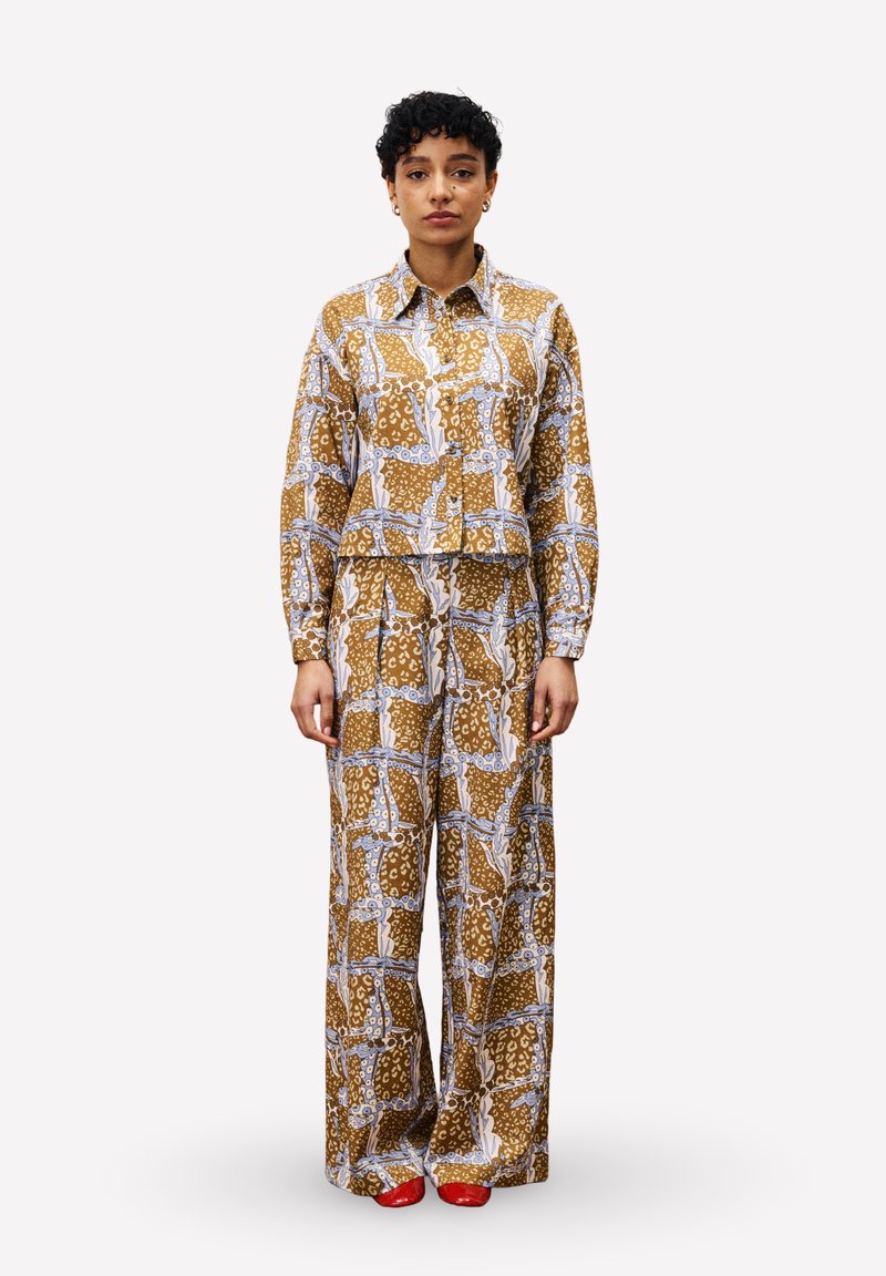 Femme debout portant une chemise à manches longues à motif marron et un pantalon large, associé à des chaussures rouges, sur un fond blanc uni.